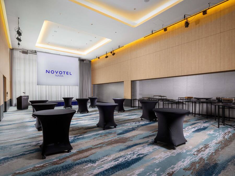 Une salle de conférence moderne dans un hôtel, dotée d’un grand écran affichant le texte « NOVOTEL NARA », de tables rondes et d’un pupitre.