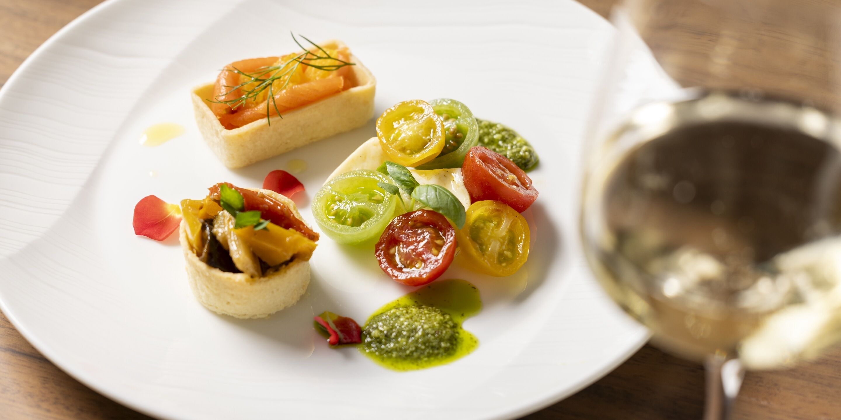 A white plate with a variety of food items including a tart, cherry tomatoes, and a glass of white wine.