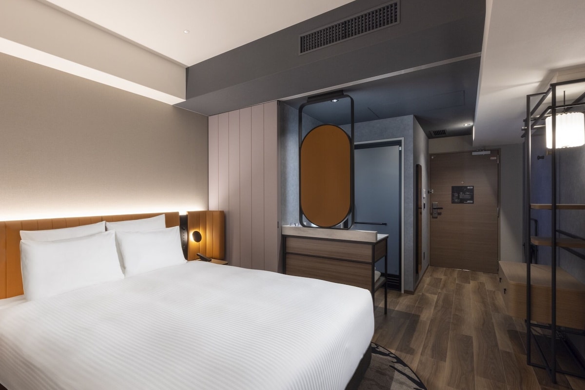 A modern hotel room with a large bed, bedside tables, a vanity with a mirror, and a walk-in closet.