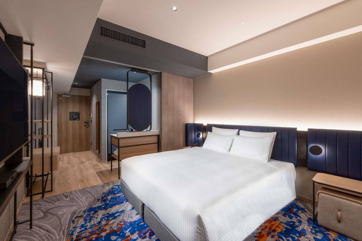 A modern hotel room with a large bed, a TV, a desk with a mirror, and a wardrobe.