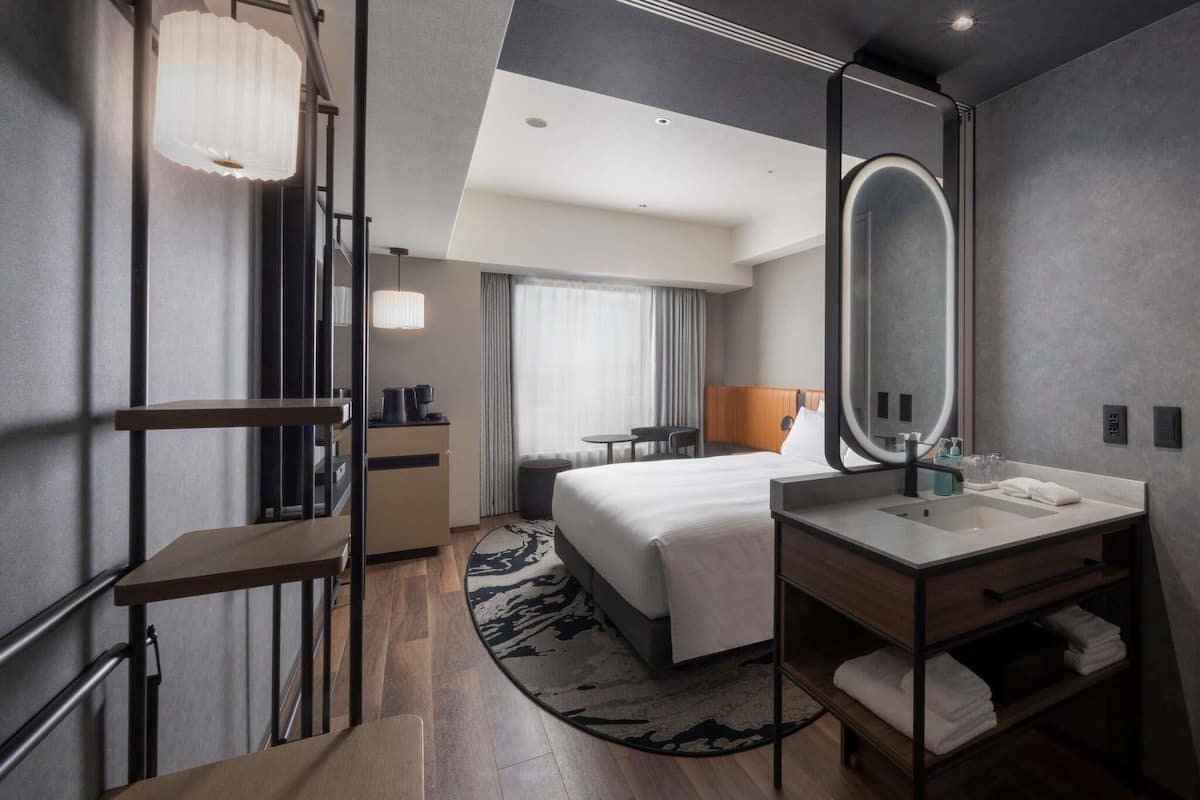 A modern hotel room with a bed, a desk, a chair, a large mirror, a sink, and a window with curtains.