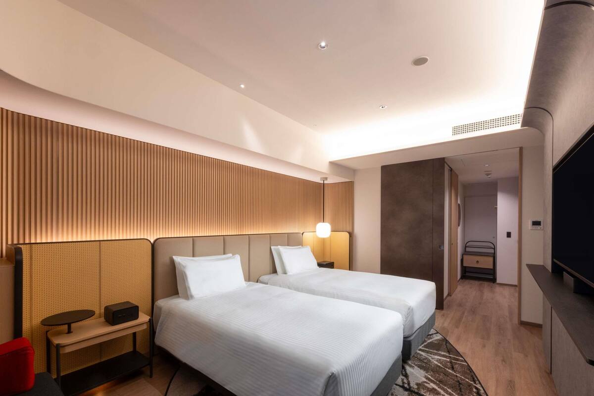 A modern hotel room with two beds, a TV, a desk, and a walk-in closet.