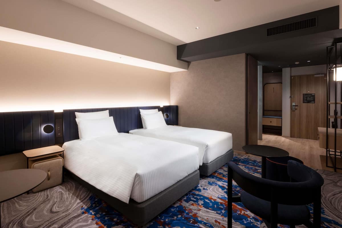 A hotel room with two beds, a small table, a chair, and a bathroom visible through a glass door.