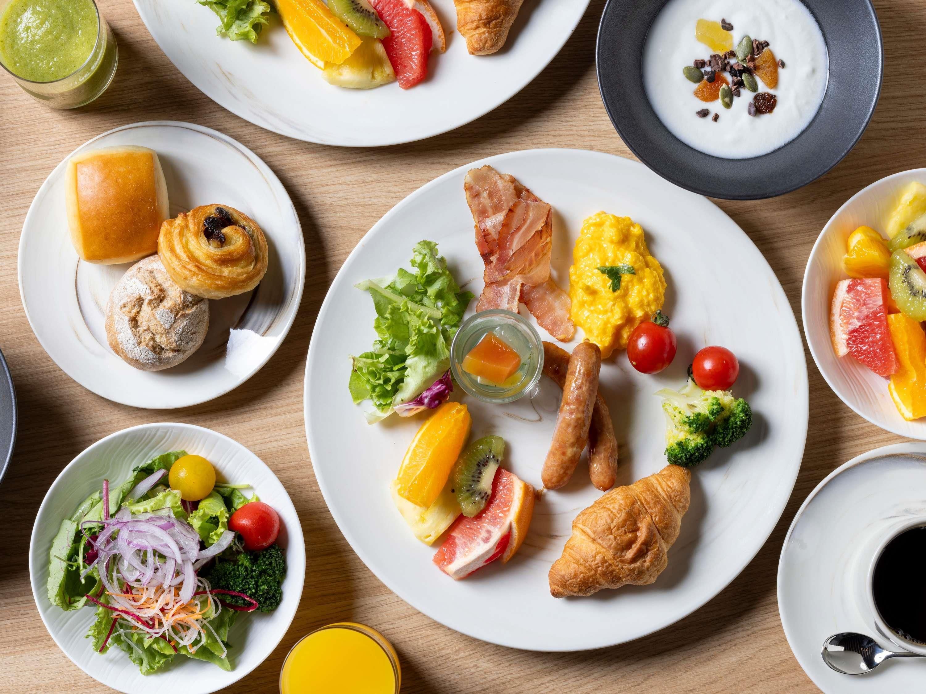 A variety of breakfast dishes including eggs, bacon, sausages, croissants, and fresh fruits on plates.