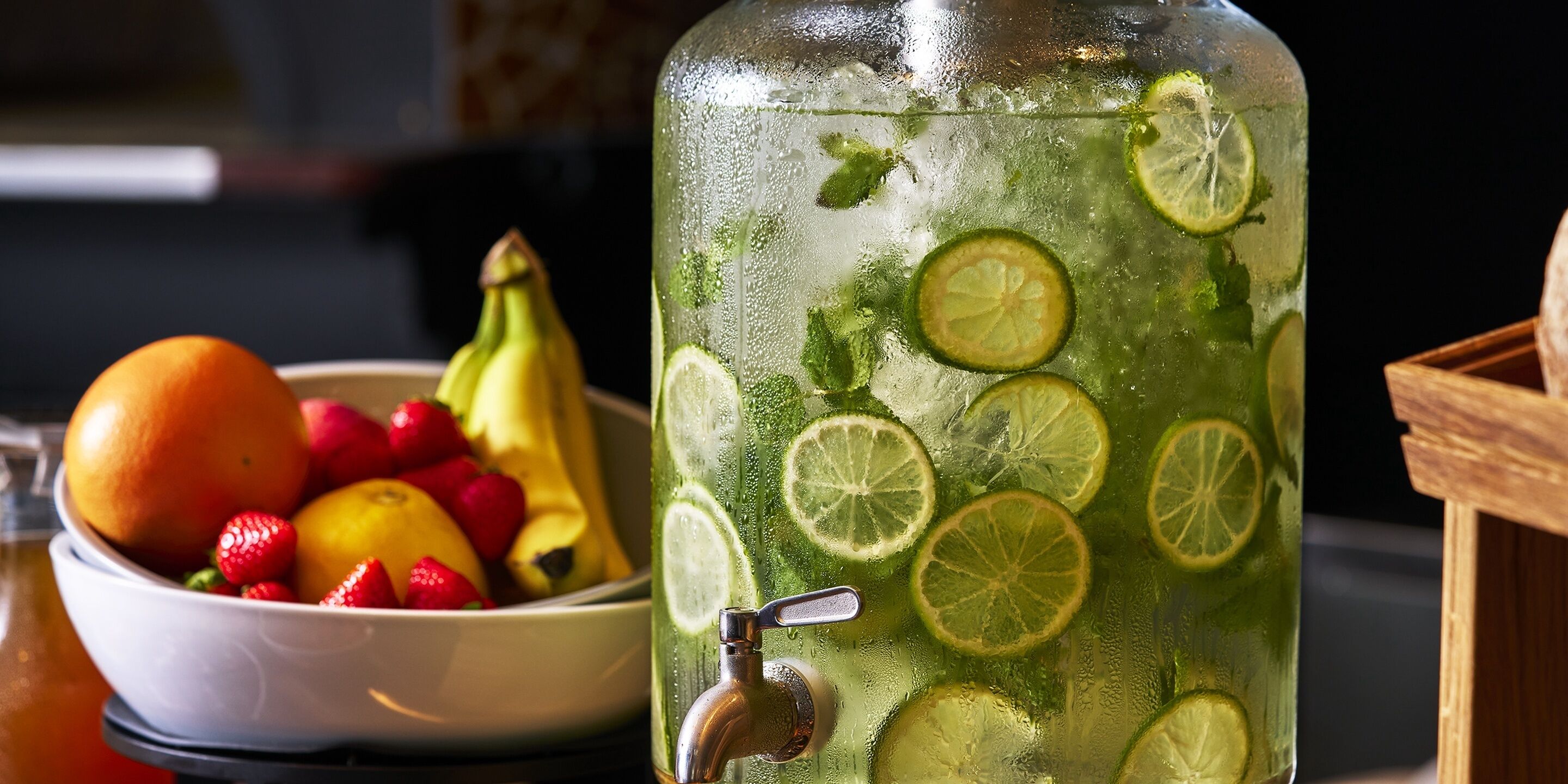 Une grande carafe en verre remplie d’une boisson verte, décorée de rondelles de citron vert, à côté d’un saladier de fruits frais.