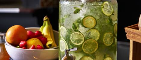 A large glass pitcher filled with a green beverage, garnished with lime slices, next to a bowl of fresh fruit.