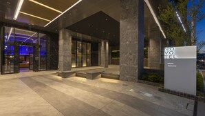 Una entrada de hotel moderna con un letrero atractivo, amplias columnas de piedra y un interior bien iluminado visible a través de las puertas de cristal.