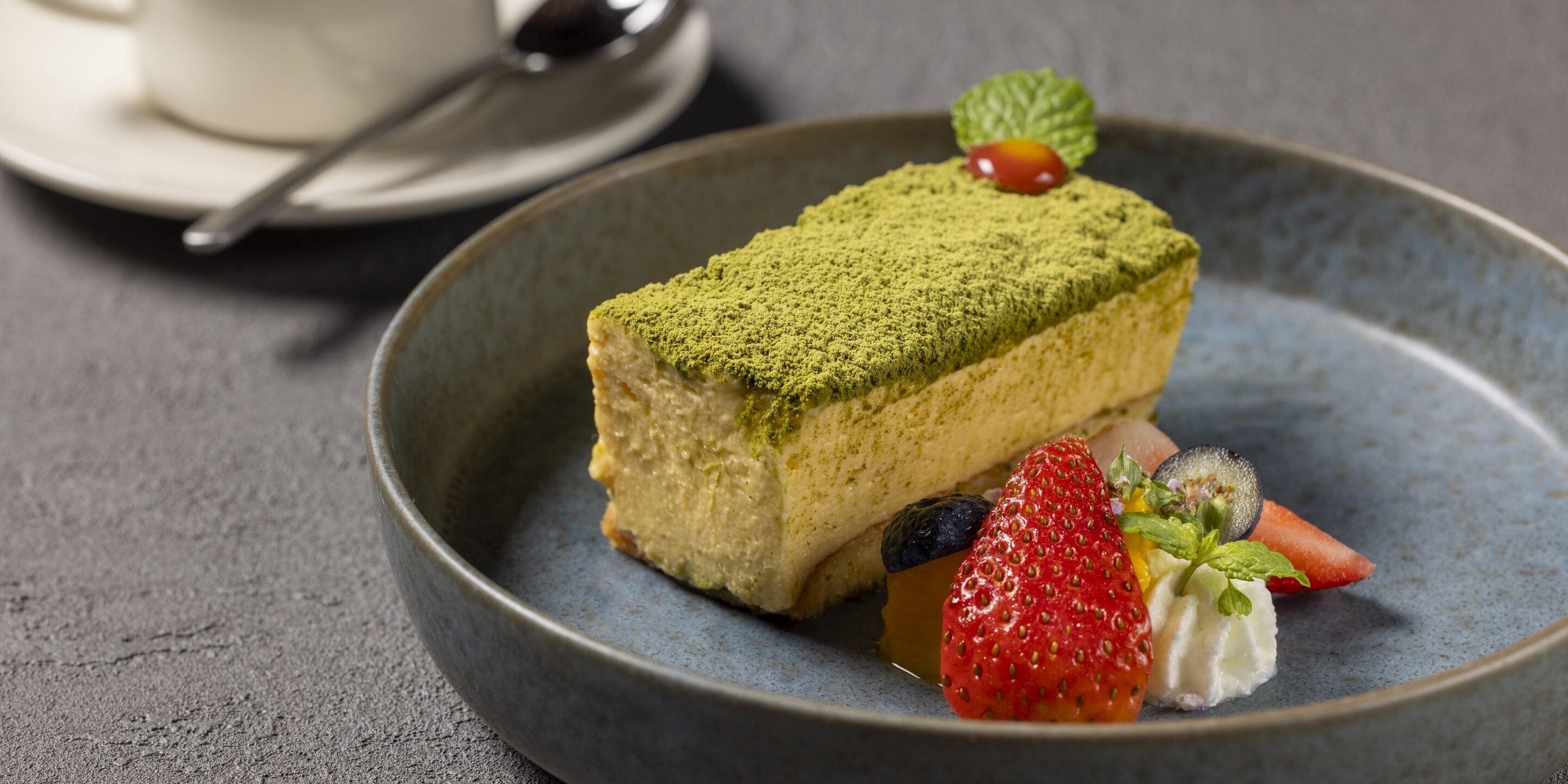 A dessert plate with a slice of cake topped with green powder, accompanied by a strawberry, kiwi, and a mint leaf.