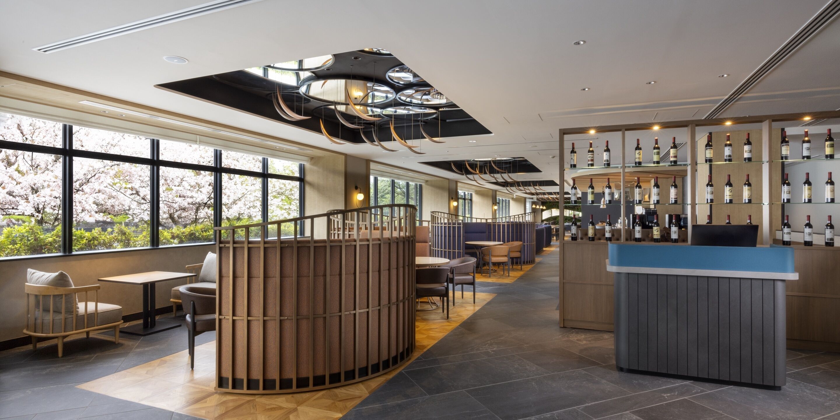 A modern hotel lobby with a curved seating area, a bar with wine bottles, and large windows offering a view of cherry blossoms.