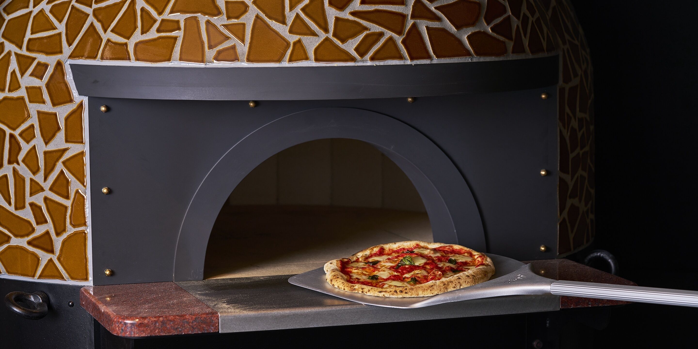 A pizza oven with a unique mosaic design, a pizza on a metal tray, and a marble countertop.