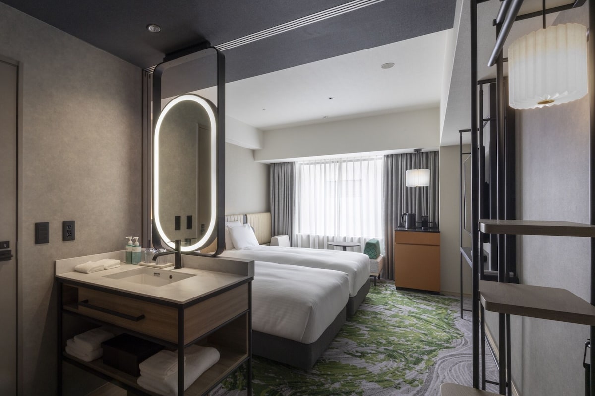 A hotel room with a large mirror, a sink, two beds, a small table, and a wardrobe.