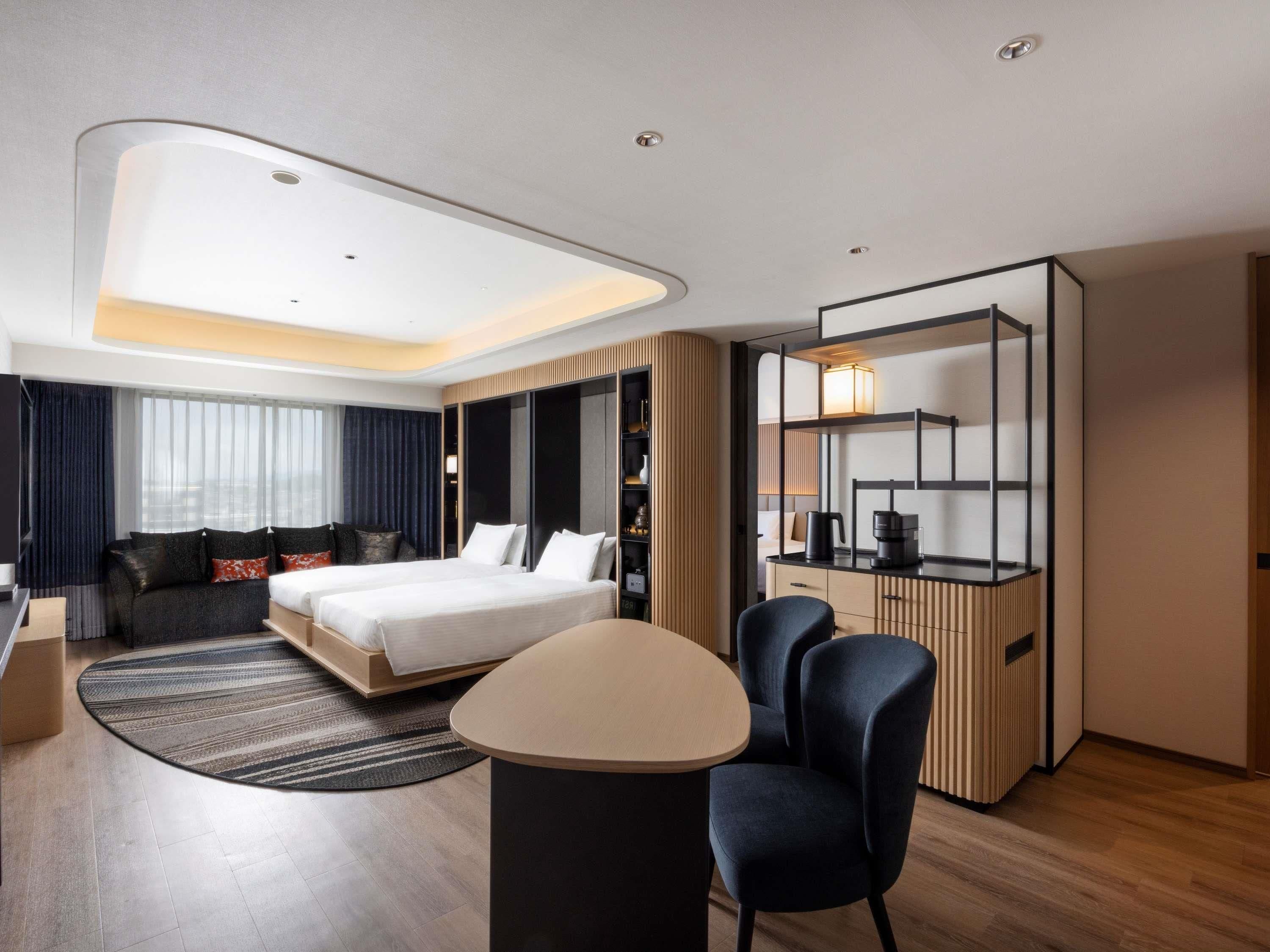 A modern hotel room with a bed, a desk, a chair, a sofa, and a built-in bookshelf.