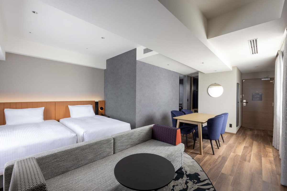 A hotel room with a bed, a sofa, a small table, a dining table with chairs, and a door.