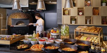 Un chef preparando comida en una cocina con horno de leña, diferentes puestos de comida y un bufé con una variedad de platos y salsas.