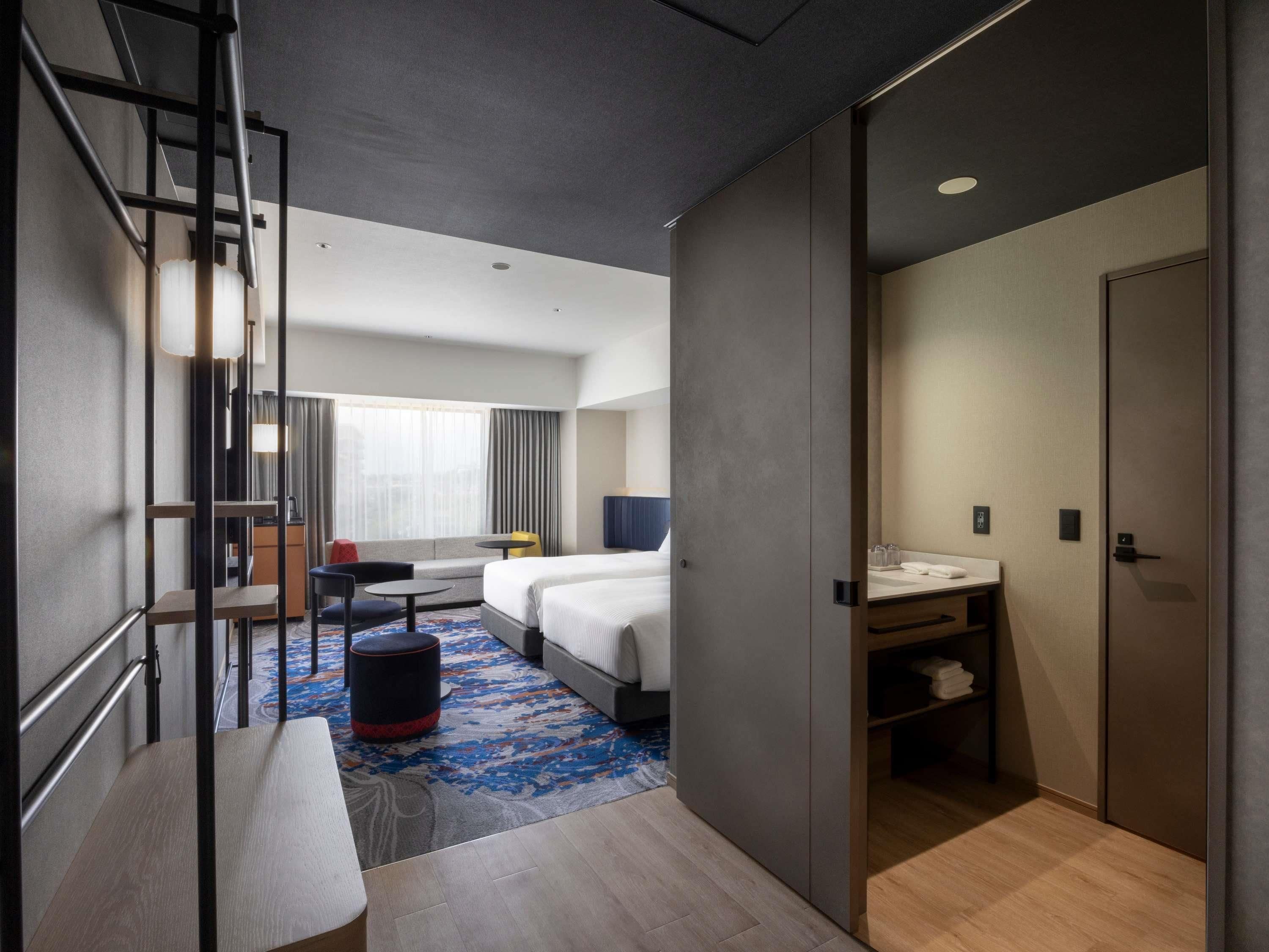 Una habitación de hotel moderna con cama, escritorio, silla, mesita, sofá y baño con lavabo y espejo.