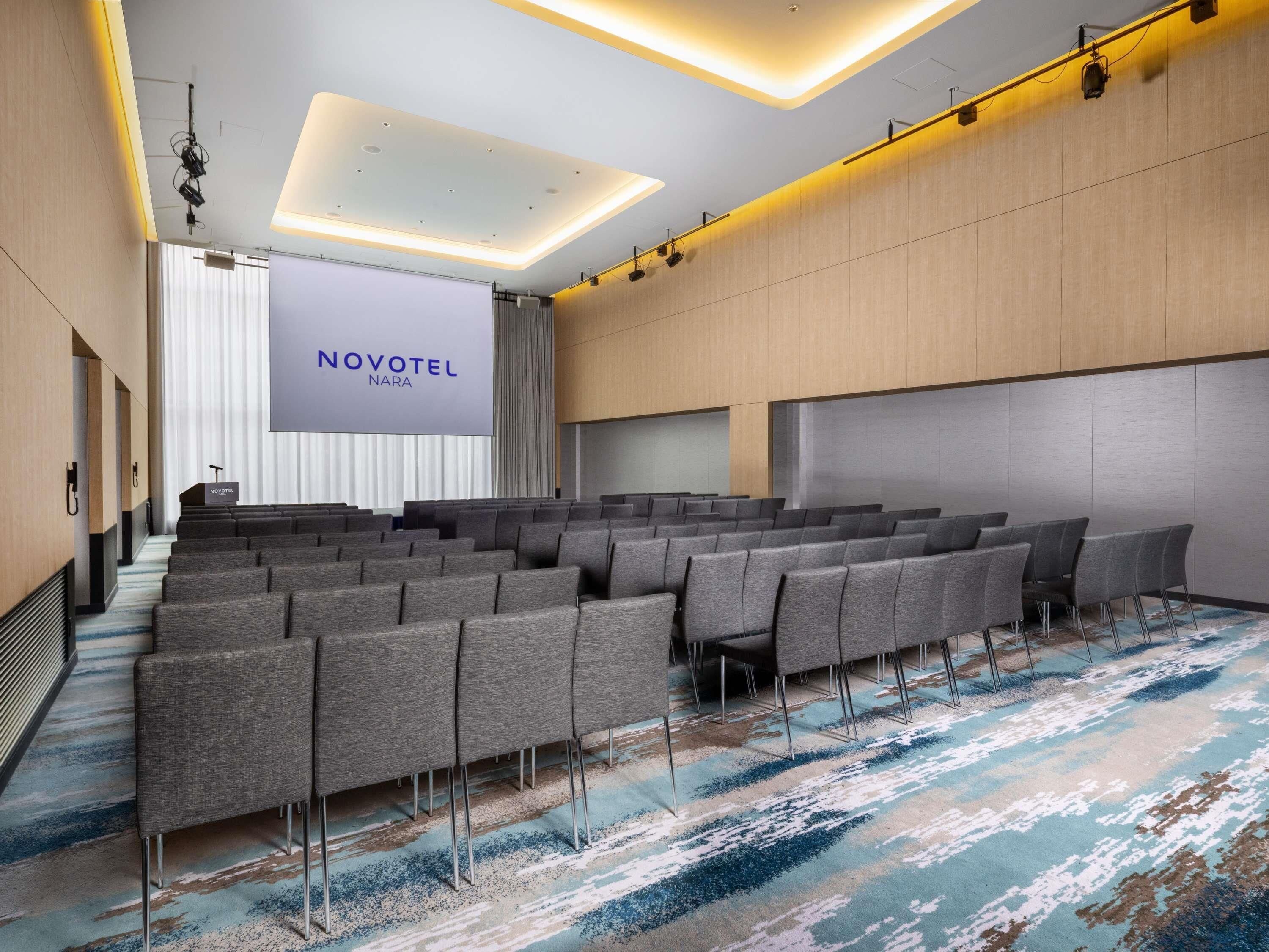 A conference room with rows of grey chairs, a large screen displaying 'NOVOTEL NARA', and a modern ceiling design.