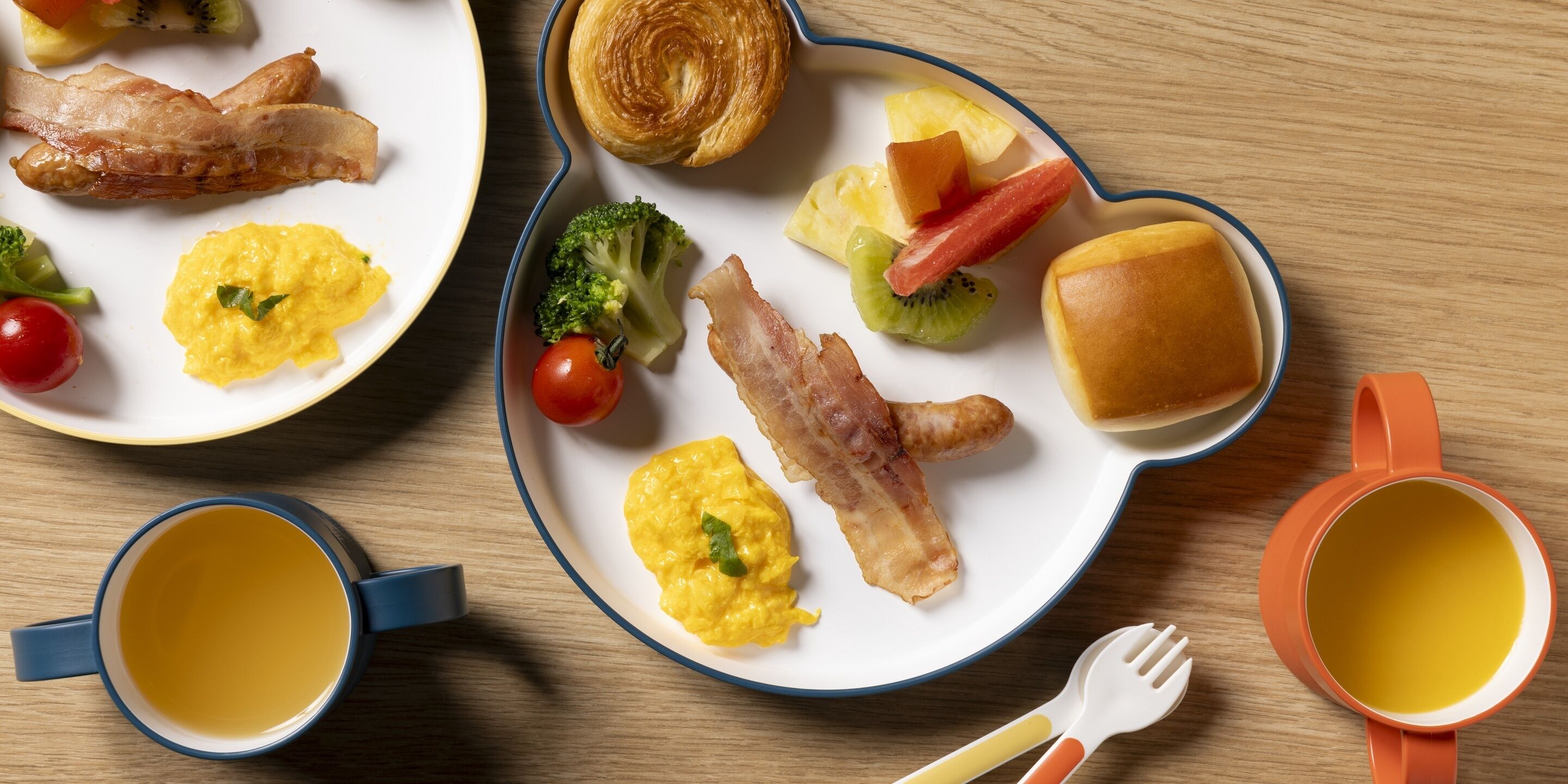 Dos platos con alimentos para el desayuno, incluyendo huevos, tocino, tostadas y fruta, sobre una mesa de madera.