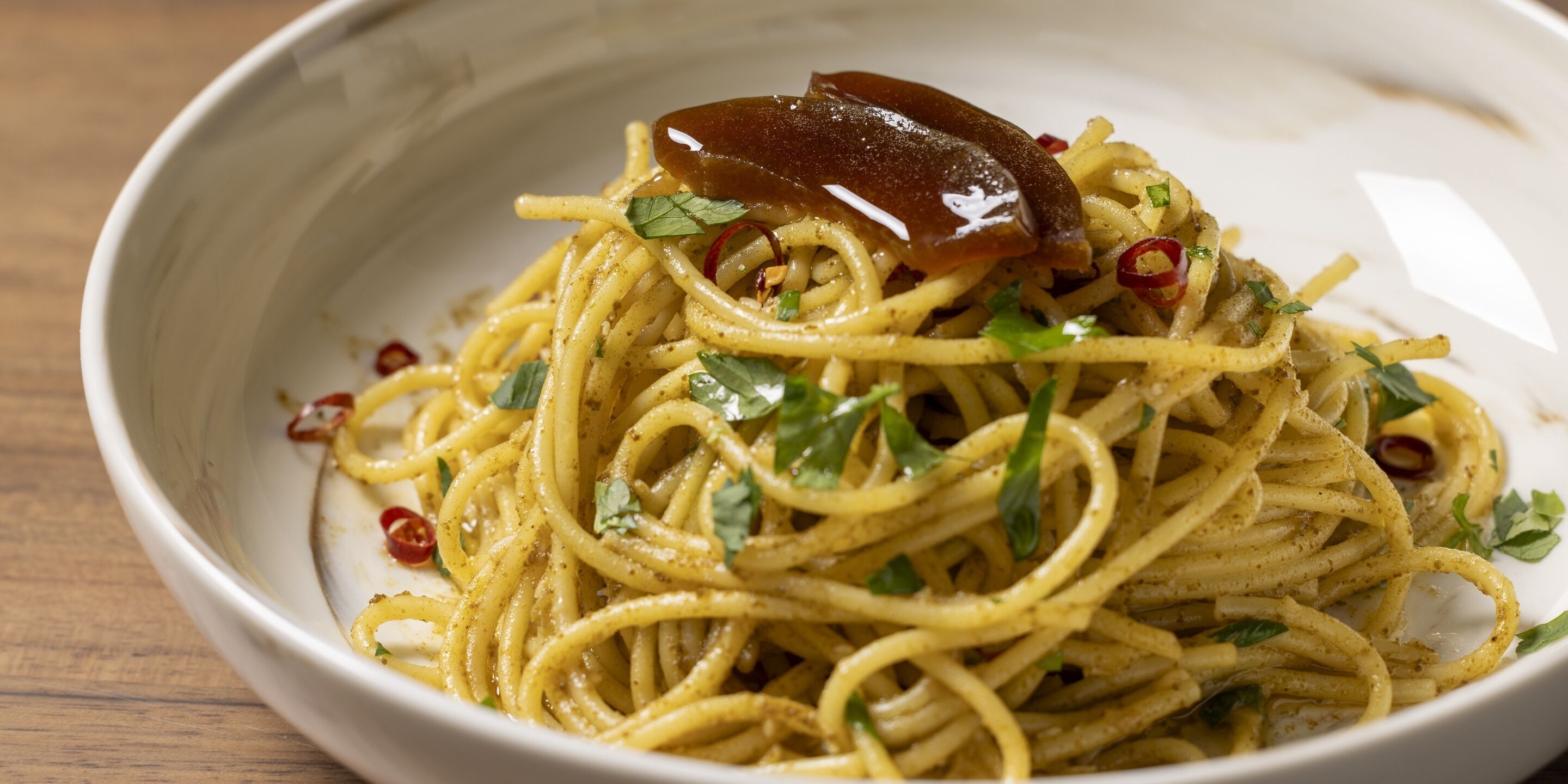 A bowl of spaghetti with a glossy topping, garnished with herbs and chili flakes.