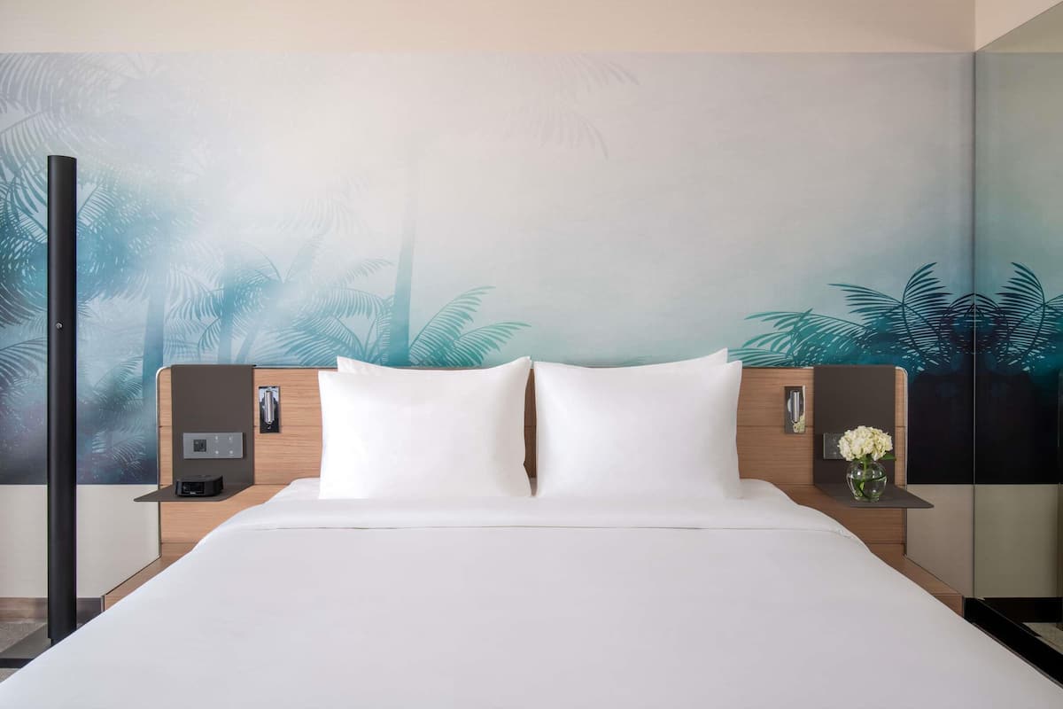 Ein Hotelzimmer mit einem groĂen Bett, zwei Nachttischen, einem an der Wand befestigten Fernseher und einem Wandbild mit tropischen BlĂ€ttern.