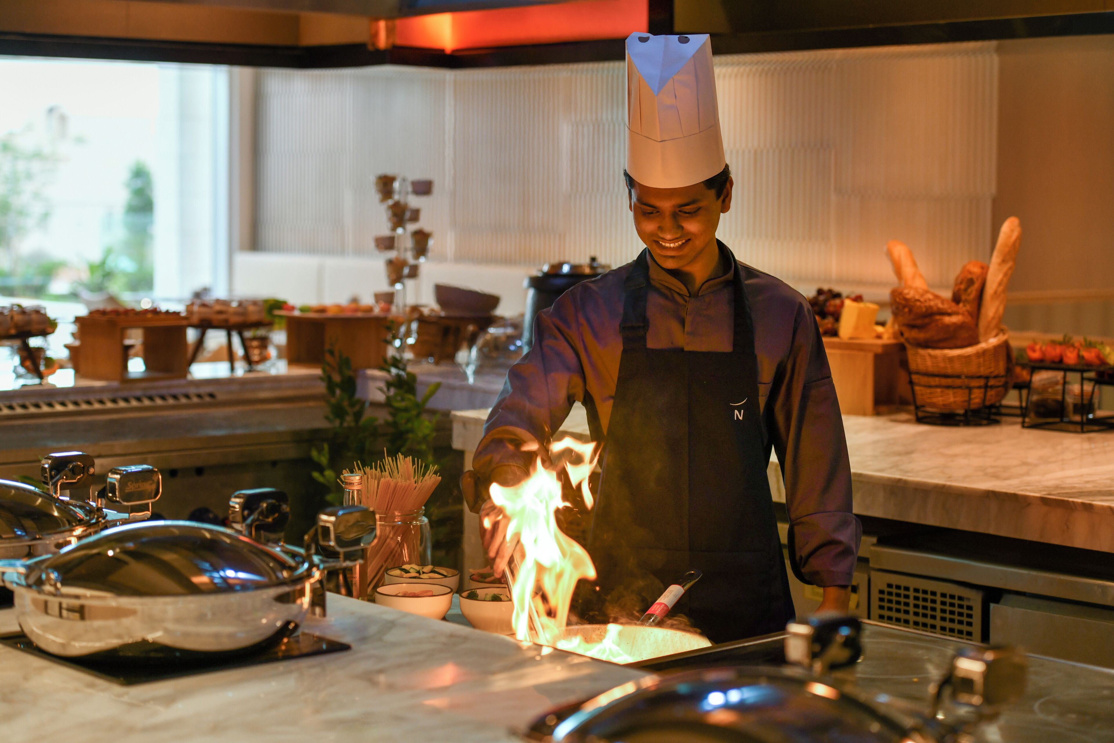 A chef in a professional kitchen with stainless steel appliances, a large window, and a variety of cooking utensils and ingredients on the counter.