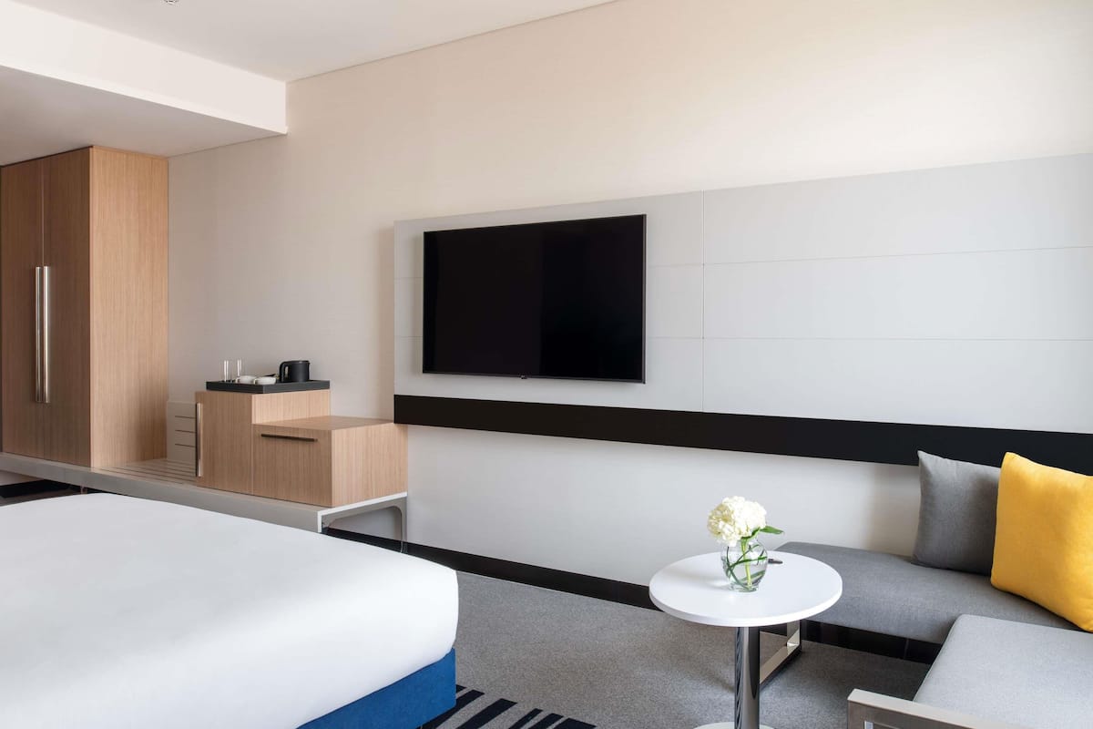 Una habitación de hotel moderna con cama, sofá, una mesita con un florero, televisor y minibar.