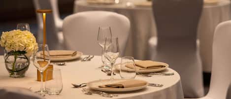 Mesa lista con platos, cubiertos y vasos, rodeada de sillas blancas.