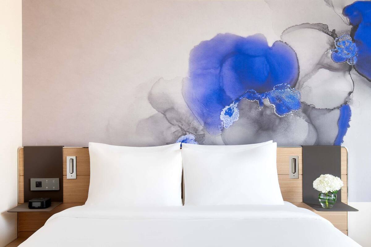 Habitación de hotel con una cama, almohadillas blancas, una mesita de noche con un reloj, un florero con flores y una pared con un cuadro abstracto floral azul.