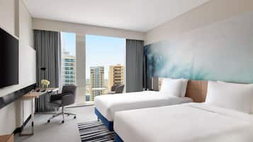 Ein Hotelzimmer mit zwei Betten, einem Schreibtisch mit Stuhl, einem Fernseher und einem großen Fenster mit Blick auf die Stadt.