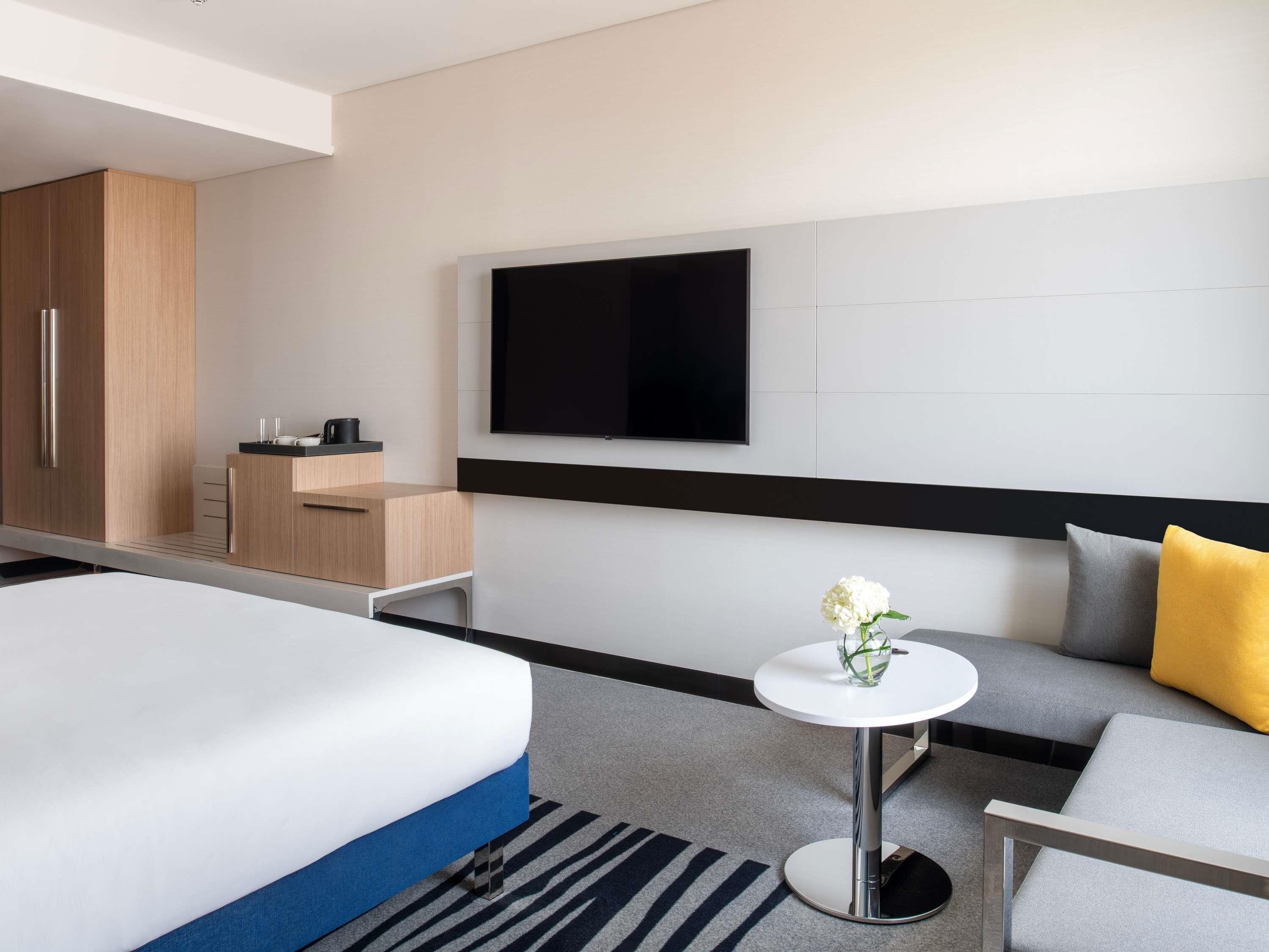 A modern hotel room with a bed, a sofa, a small table with a vase, a TV mounted on the wall, and a wooden cabinet.