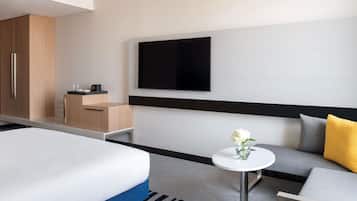 A modern hotel room with a bed, a sofa, a small table with a vase, a TV mounted on the wall, and a wooden cabinet.