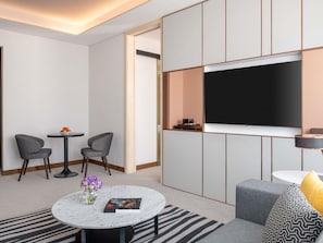 Moderni hotellihuone, jossa on litteänäyttöinen televisio, harmaalla kankaalla verhoiltu istuin, marmorinen sohvapöytä ja pieni sivupöytä, jossa on maljakko täynnä kukkia.