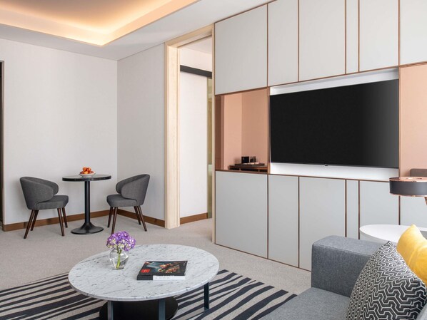 Una habitación de hotel moderna con una televisión de pantalla plana, asientos tapizados en gris, una mesita de centro de mármol y una mesita auxiliar con un jarrón de flores.