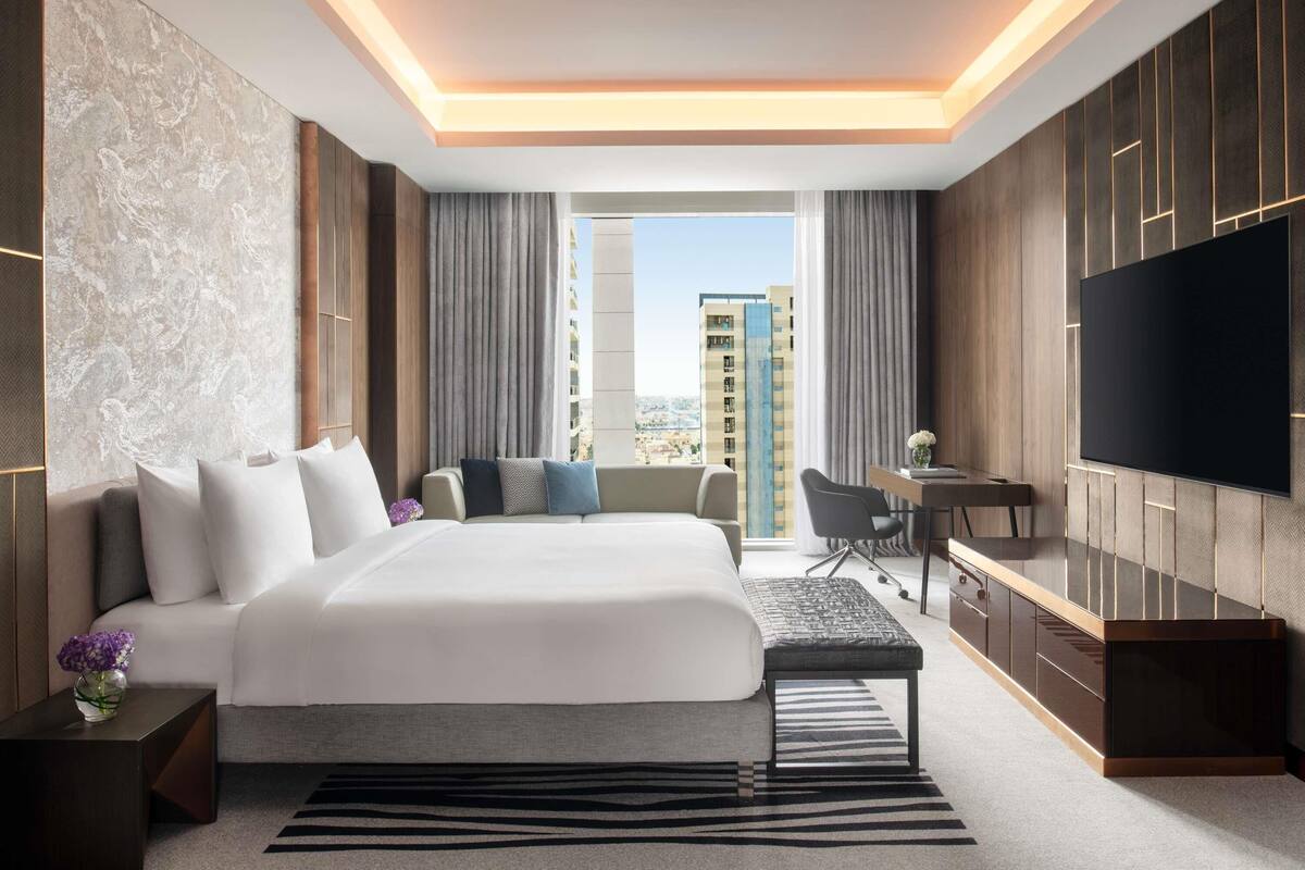 A modern hotel room with a large bed, a desk, a TV, and a view of the cityscape.