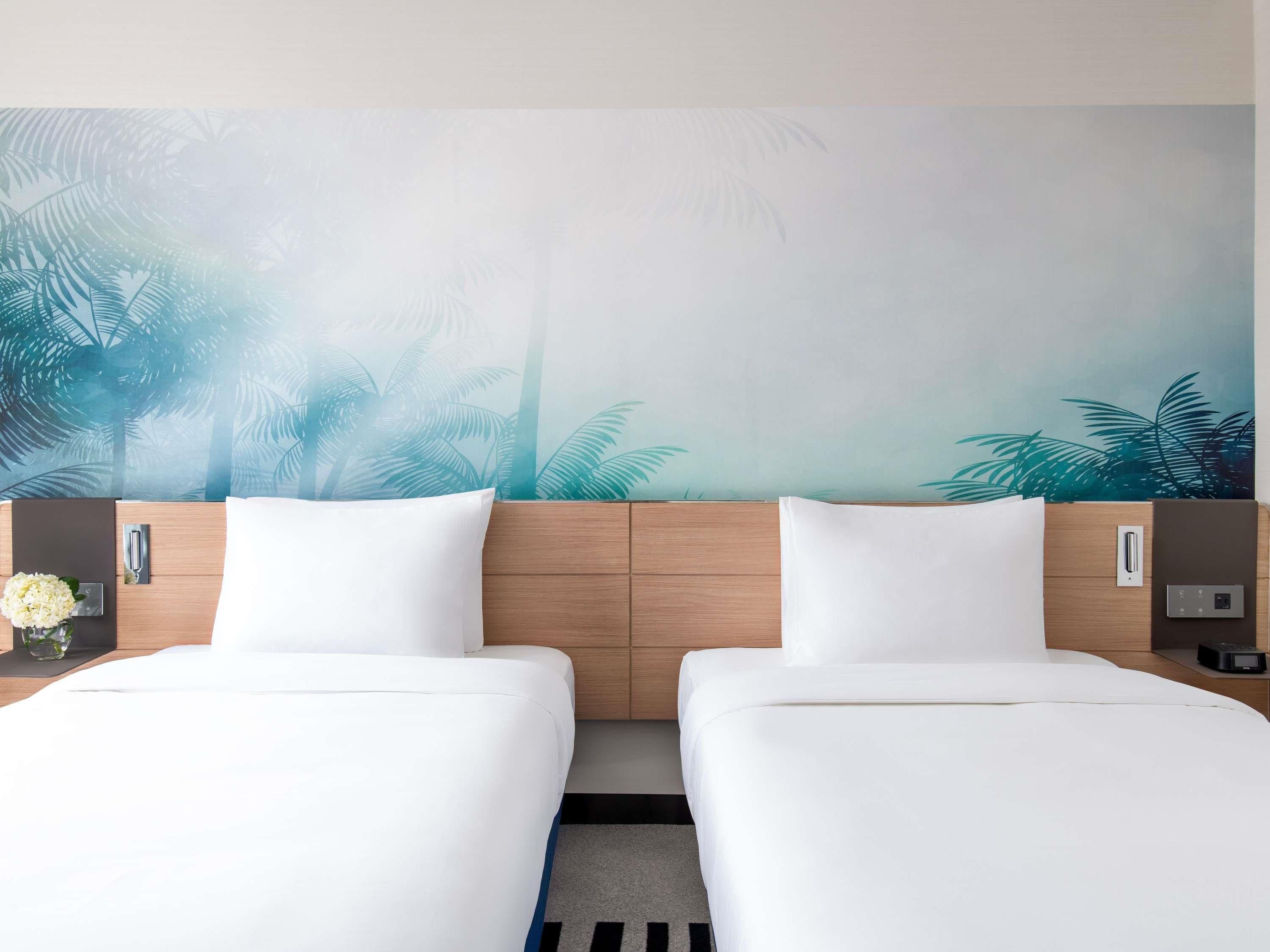 A hotel room with two beds, a wooden headboard, a wall mural of palm trees, a nightstand with a vase of flowers, and a digital clock.