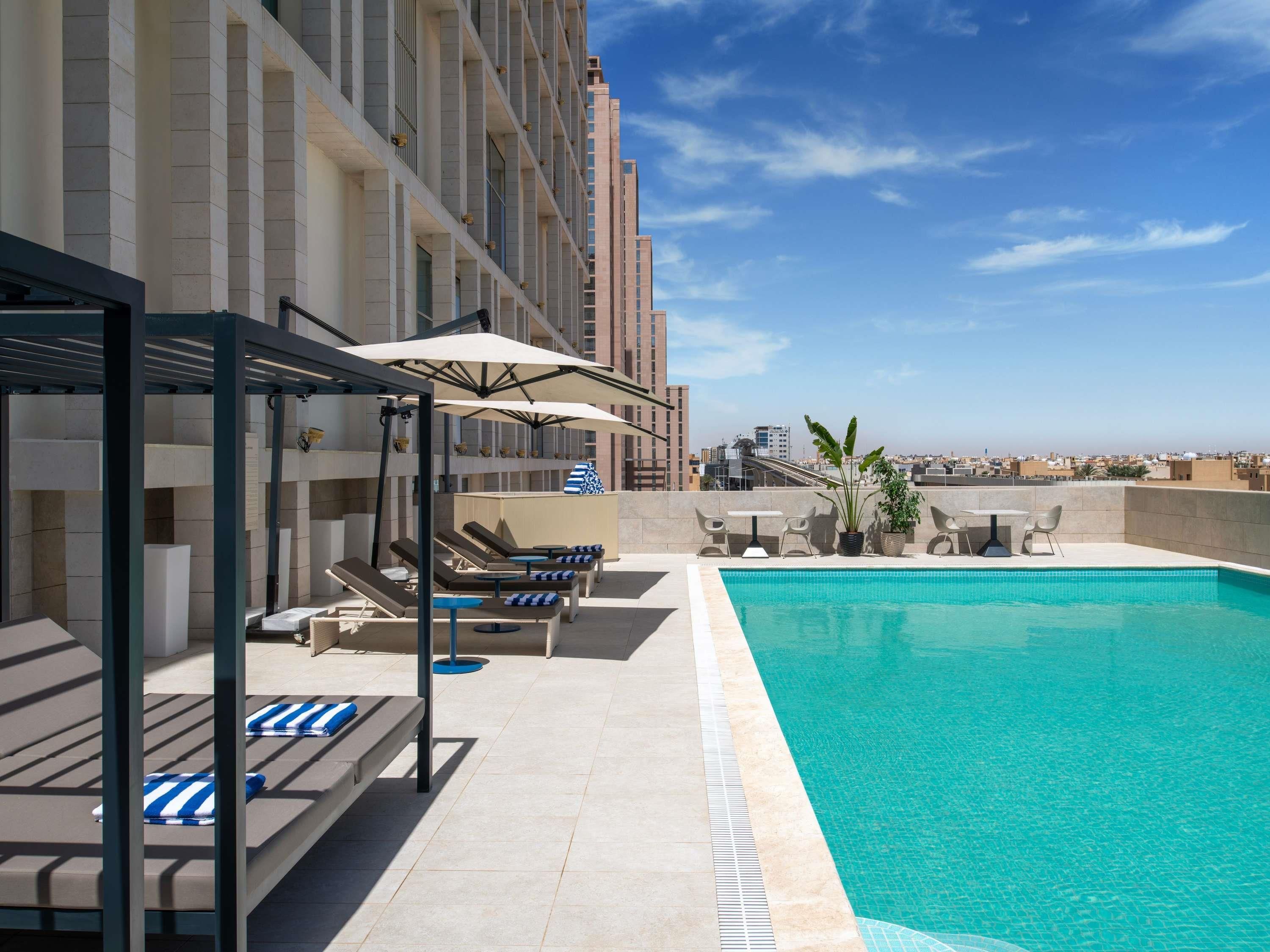 Zona de piscina en la azotea con tumbonas y sombrillas, una pequeña mesa con sillas y vistas al horizonte de la ciudad.