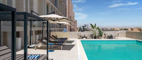 Zona de piscina en la azotea con tumbonas y sombrillas, una pequeña mesa con sillas y vistas al horizonte de la ciudad.