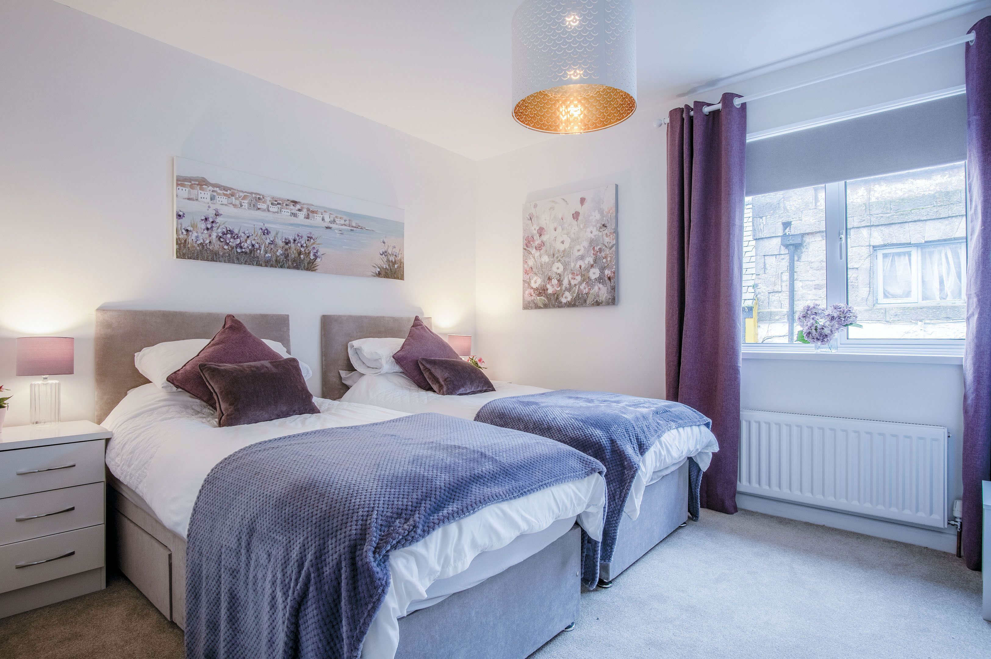 A bedroom with two beds, a nightstand, a lamp, and artwork on the walls.