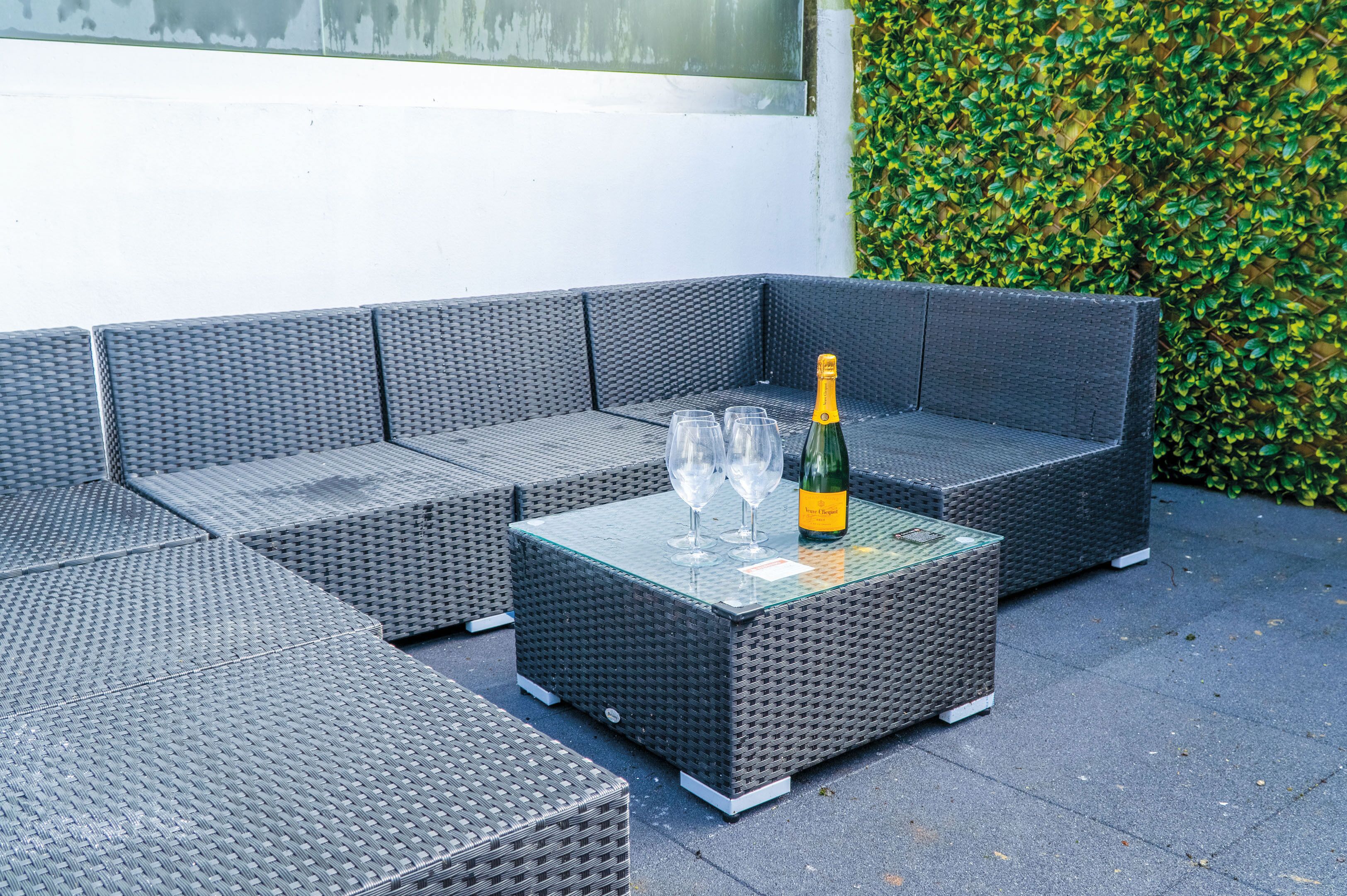 Un espace extérieur aménagé avec des sièges en tissu gris, une table avec un plateau en verre, deux verres à vin et une bouteille de champagne.