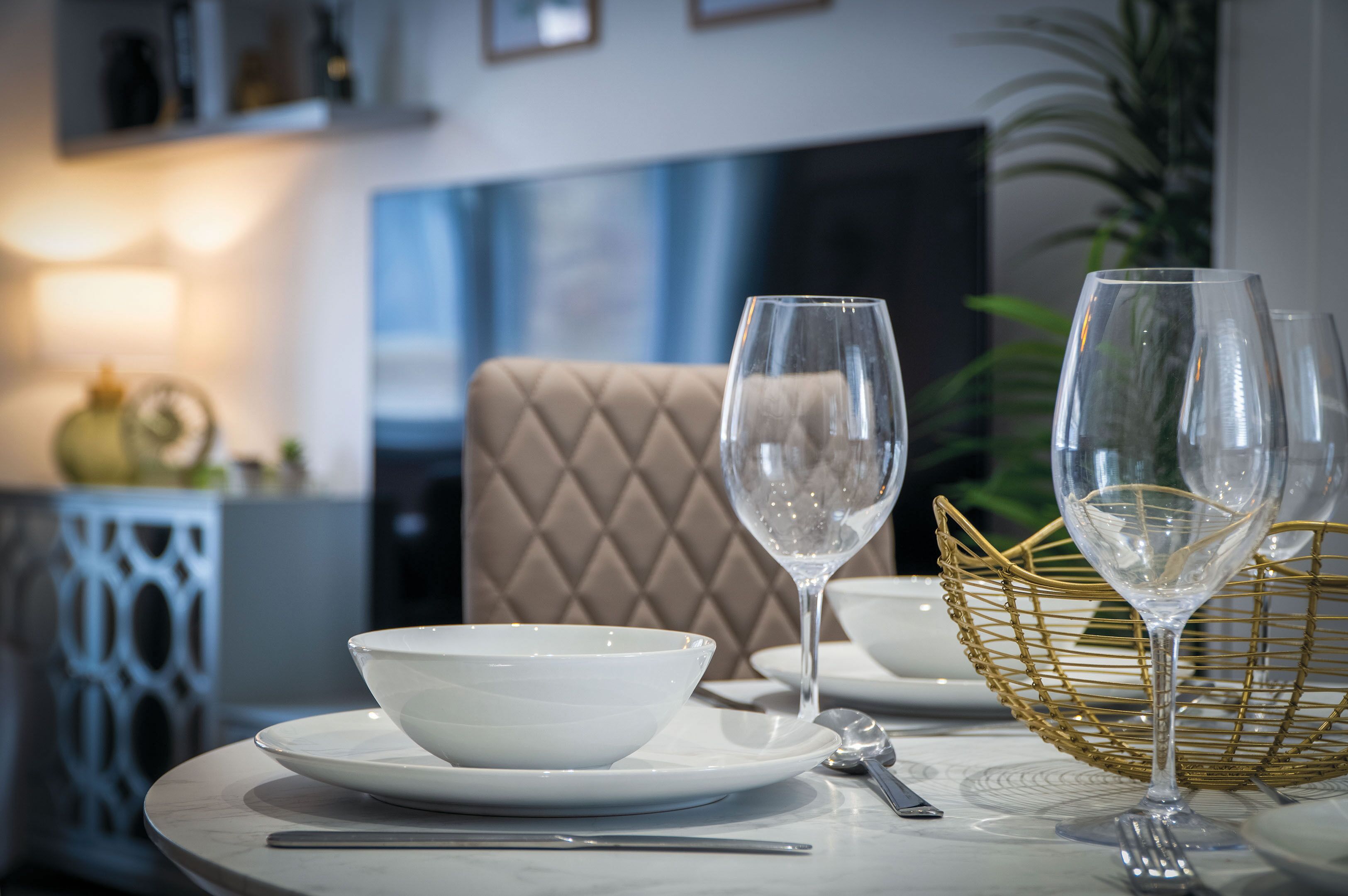 A dining table with a white bowl, two wine glasses, and a gold basket.
