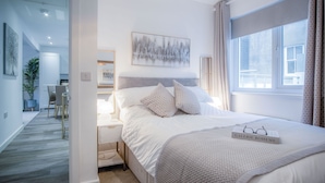 Une chambre à coucher moderne avec un lit soigneusement fait, une table de chevet, une fenêtre avec des rideaux et une vue sur l’extérieur.