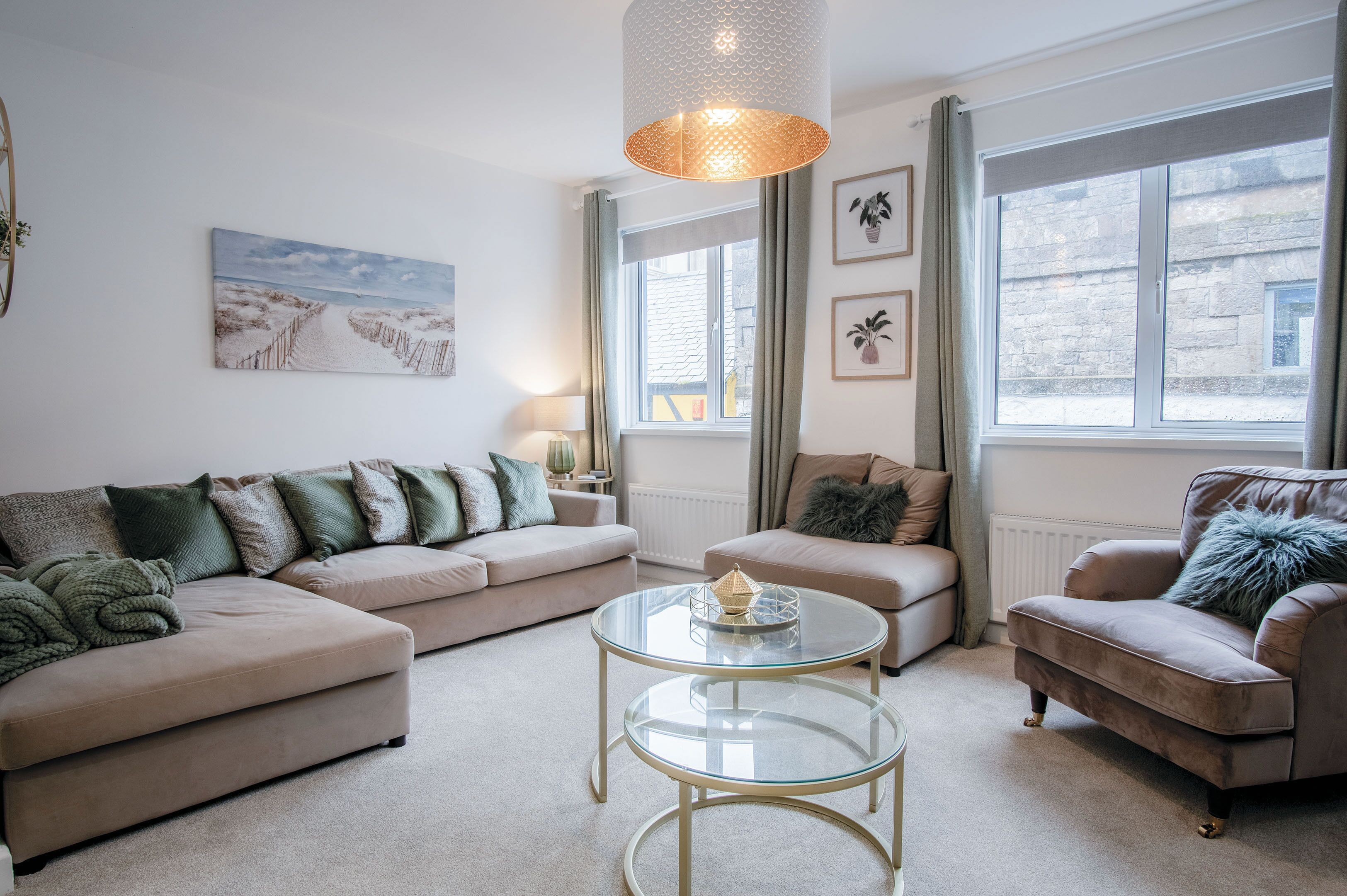 Ein Wohnzimmer mit einem beigen Sofa, einem runden Glastisch und einem beigen Sessel. Ein Fenster mit Vorhängen, eine Lampe und ein Gemälde sind an der Wand zu sehen.