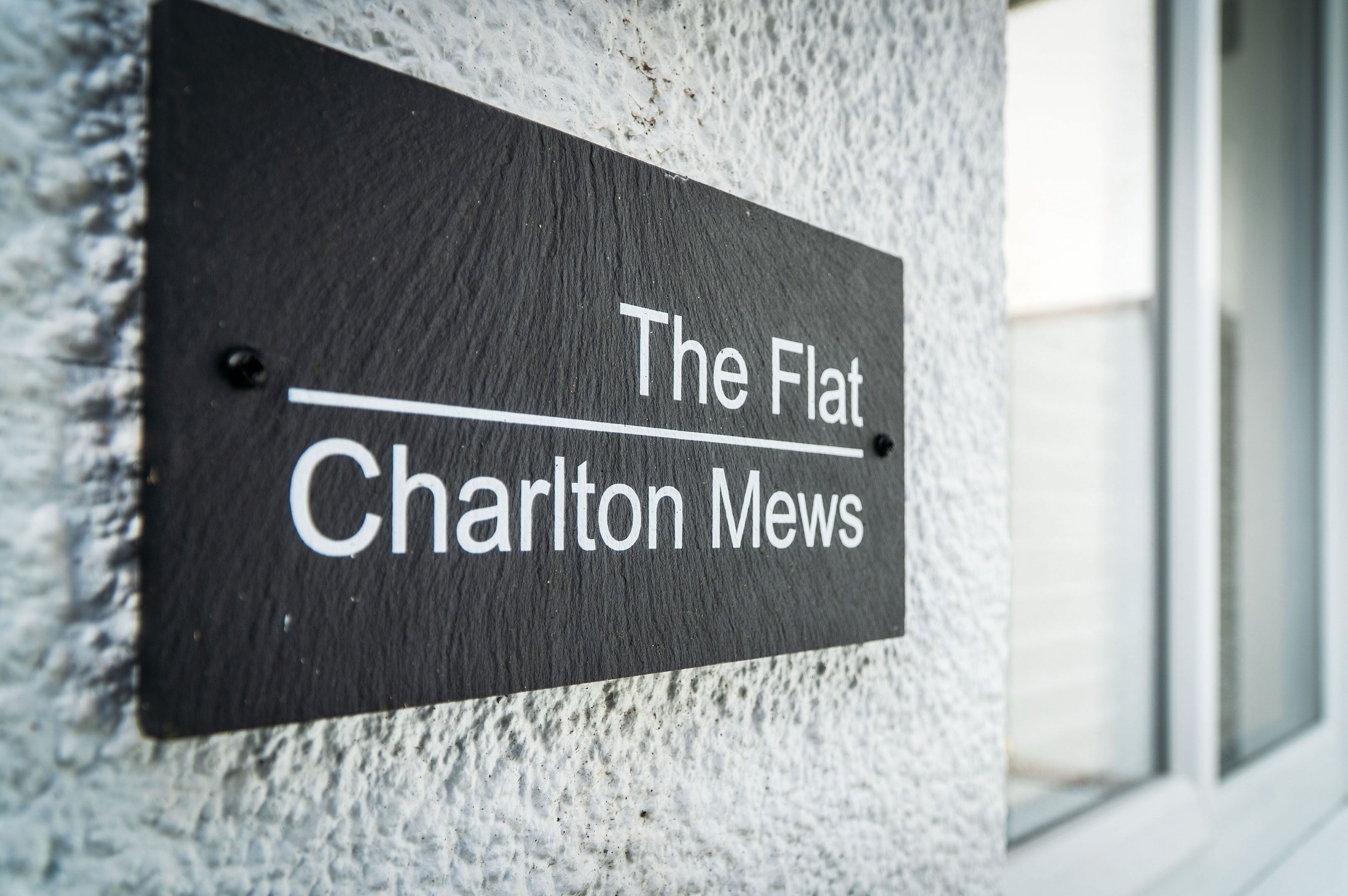 A sign with the text "The Flat Charlton Mews" mounted on a textured wall.