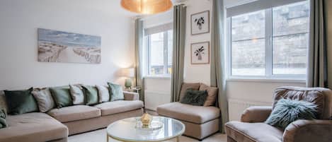Egy modern nappali, melyben bézs színű kanapé, egy kerek, üveg asztallapú dohányasztal és egy bőr fotel található. Egy nagy ablakon át fény szűrődik be, melyen függöny van, a falon egy kép, a mennyezetről pedig egy lógó lámpa világítja meg a teret.