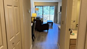Un pasillo que conduce a una sala de estar con sofá, lámpara y vistas a través de un ventanal.