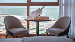 Quarto com uma grande janela com vista para a praia, uma mesa de madeira com uma garrafa de champanhe, duas cadeiras e uma cama com roupa de cama branca.