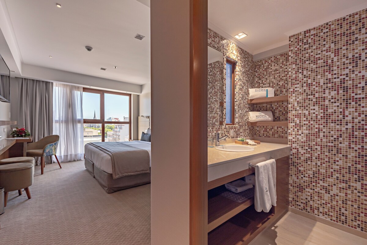 A hotel room with a large bed, a desk, a chair, a bathroom with a tiled wall, and a view of the city.