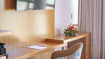 A hotel room with a wooden desk, a flat-screen TV mounted on the wall, a chair with a blue and white pattern, and a window with curtains.