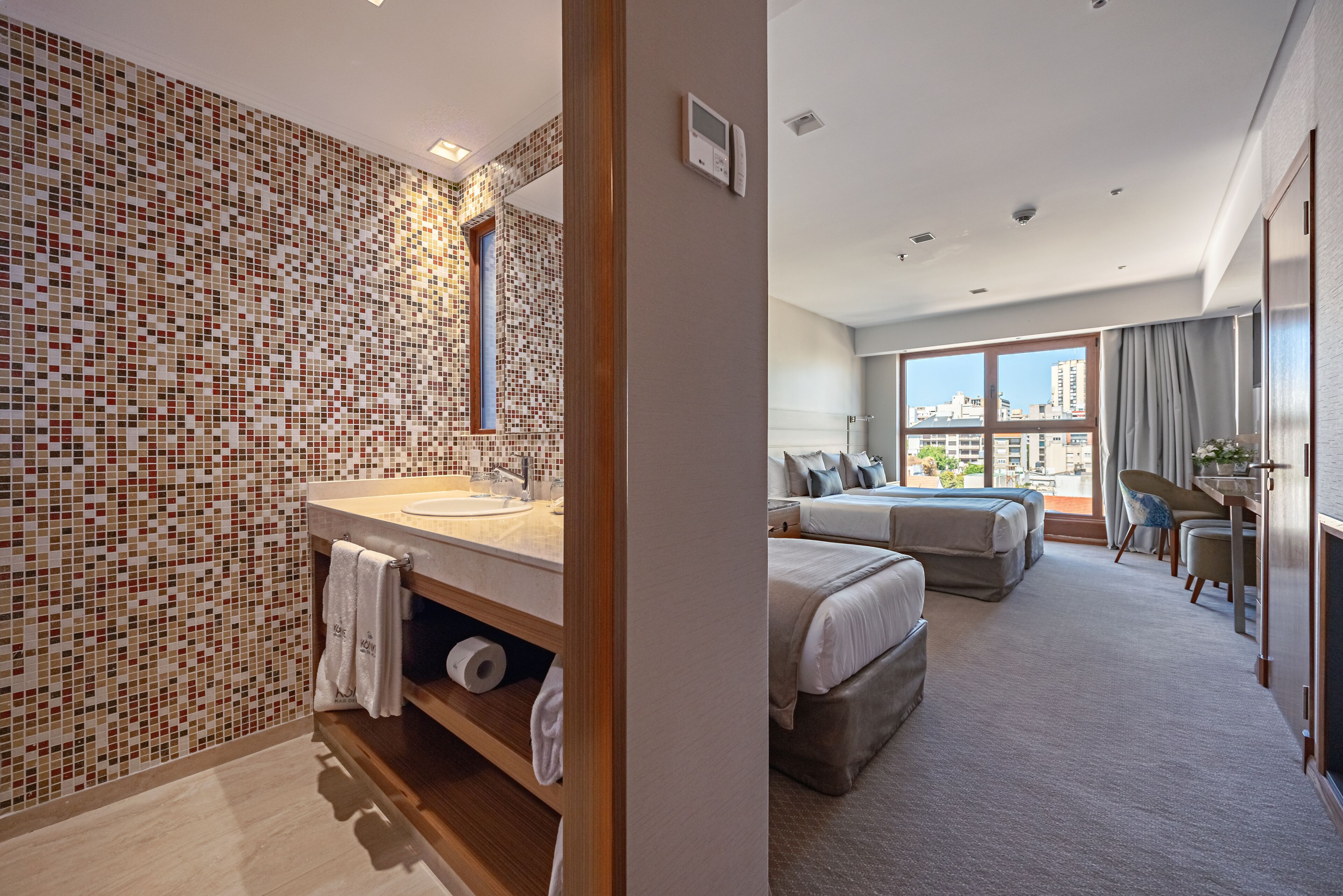 A hotel room with a bathroom featuring a mosaic tile wall, a large mirror, and a wooden vanity with a sink and towel rack. The room has two beds, a large window with a city view, and a seating area with a chair and table.