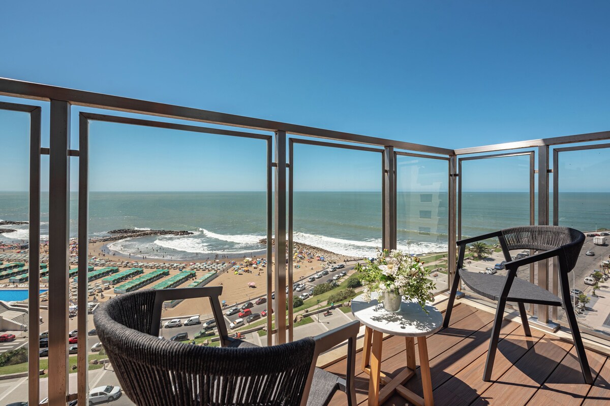 A balcony with a view of the beach and ocean. There are two chairs and a small table with a flower vase.