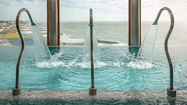 Piscina com três jatos de água, chão em mosaico e vista para o oceano através de grandes janelas.